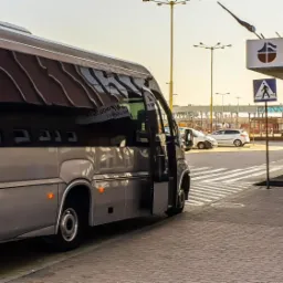 Szary bus z otwartymi drzwiami stoi przed terminalem promowym w Świnoujściu, widoczny znak przejścia dla pieszych.