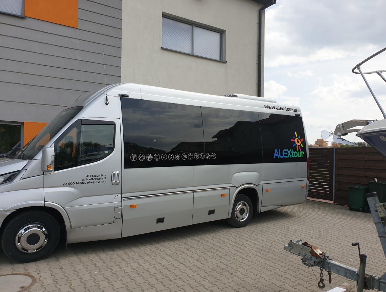 Srebrny bus turystyczny ALEXtour z przyciemnianymi szybami, zaparkowany na brukowanym podjeździe, z widocznym adresem firmy na karoserii i logiem na szybie.
