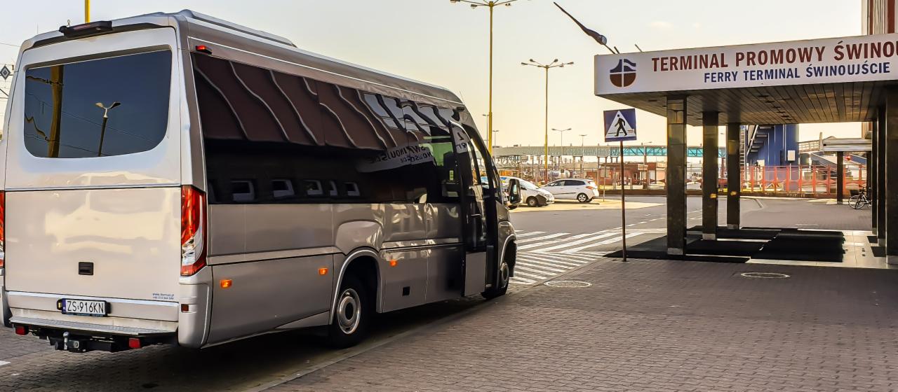 Szary bus z otwartymi drzwiami stoi przed terminalem promowym w Świnoujściu, widoczny znak przejścia dla pieszych.