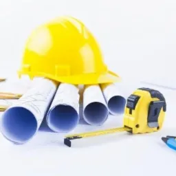 Żółty kask budowlany leży na zwiniętych planach, obok rozwinięta miarka i ołówek na białym tle.