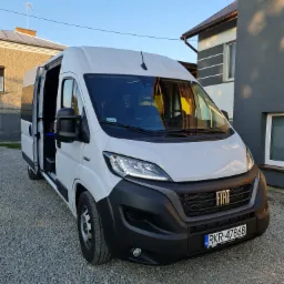 Biały bus Fiat Ducato z otwartymi drzwiami, zaparkowany na żwirowym podjeździe w Iwoniczu, widok z przodu na tle budynku.