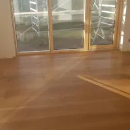 Jasny parkiet ułożony w pokoju z dużym oknem, za którym widoczne rusztowanie i zieleń ogrodu.