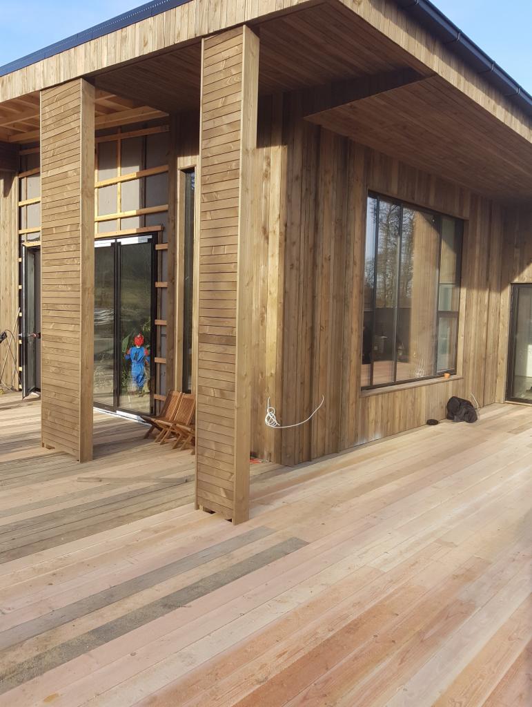 Drewniana elewacja domu z dużymi oknami i tarasem z desek; widoczny pracownik w kombinezonie wewnątrz budynku.