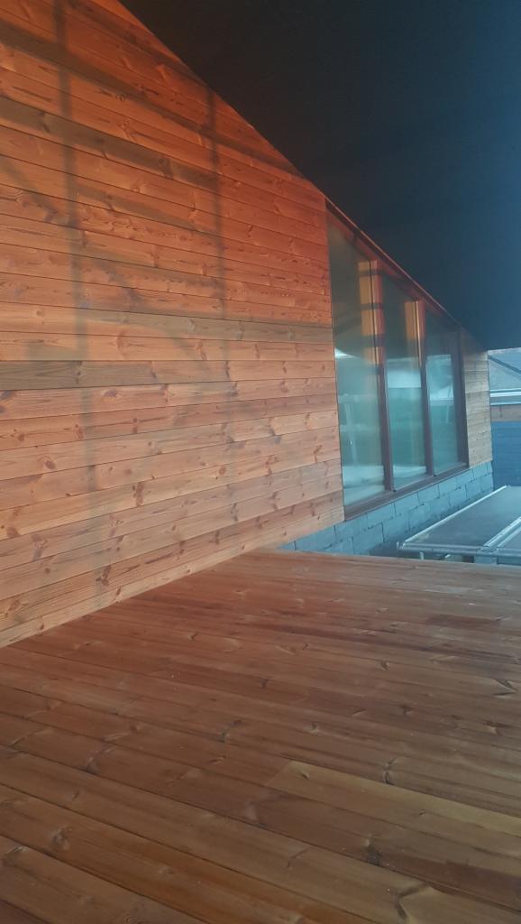 Drewniana elewacja budynku z dużymi oknami i drewnianym tarasem, widoczne słońce odbijające się od powierzchni drewna.