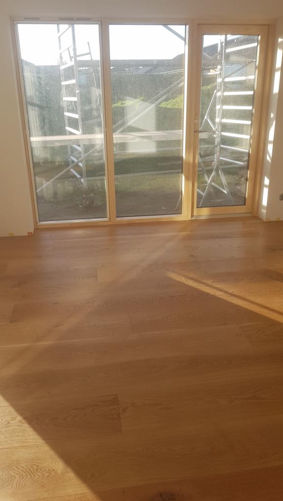 Jasny parkiet ułożony w pokoju z dużym oknem, za którym widoczne rusztowanie i zieleń ogrodu.