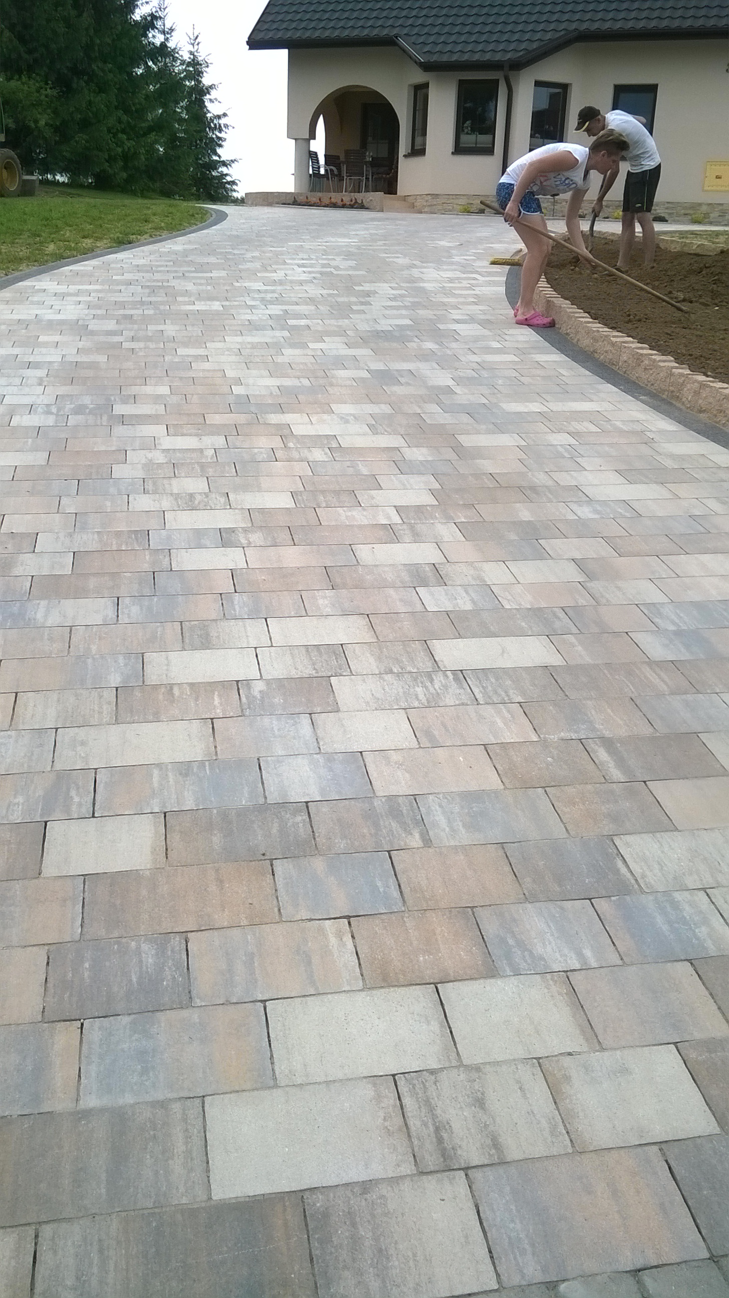 Szeroki podjazd z ułożonej kostki brukowej w odcieniach szarości i beżu, z widocznymi pracownikami wyrównującymi ziemię wzdłuż obrzeża, przed domem z kolumnami.