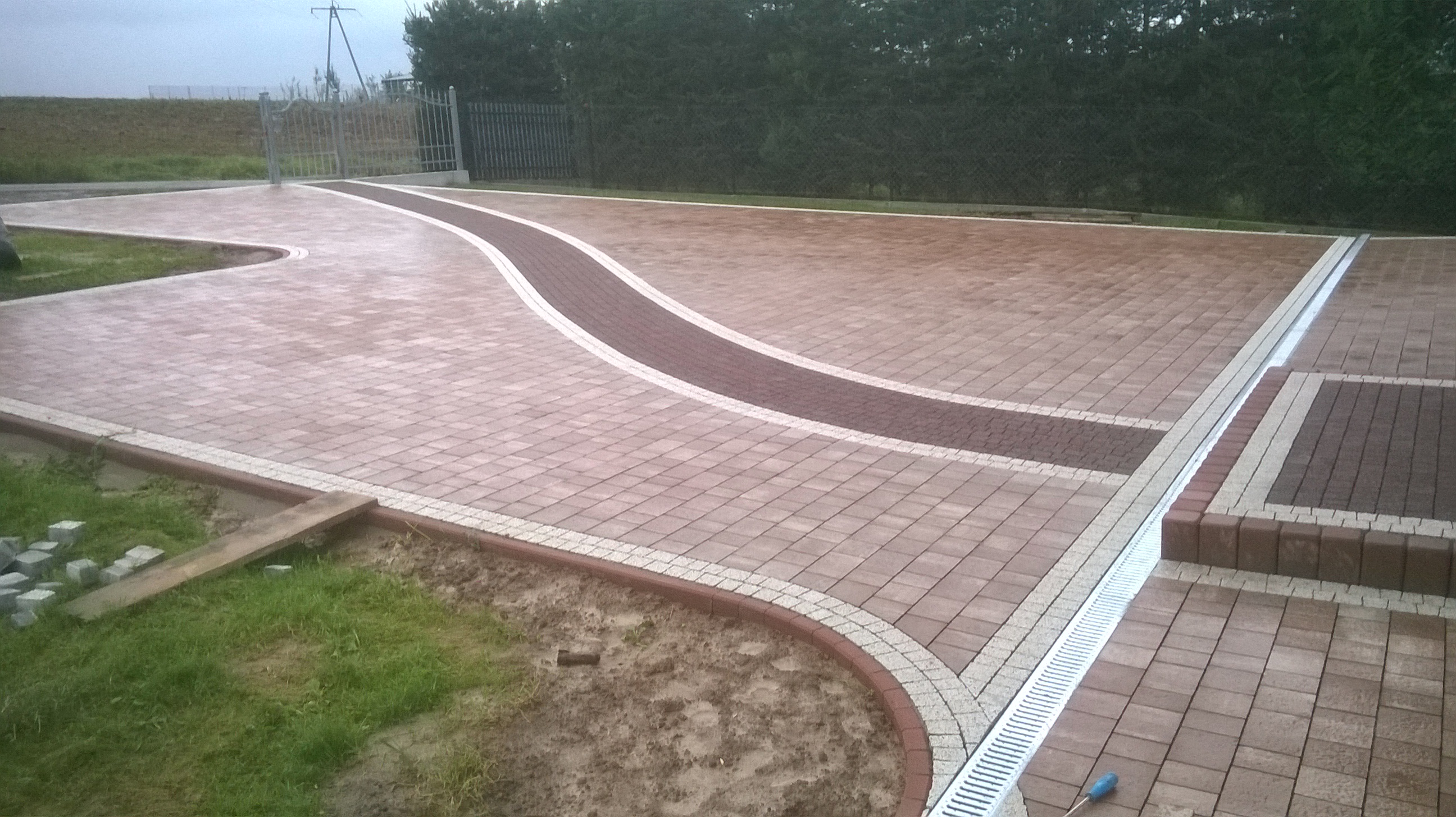 Ukończona nawierzchnia z kostki brukowej w odcieniach brązu i beżu, z dekoracyjnym wzorem w kształcie fali i systemem odwodnienia liniowego. Widoczne obrzeża z kostki, trawnik i częściowo ułożone...