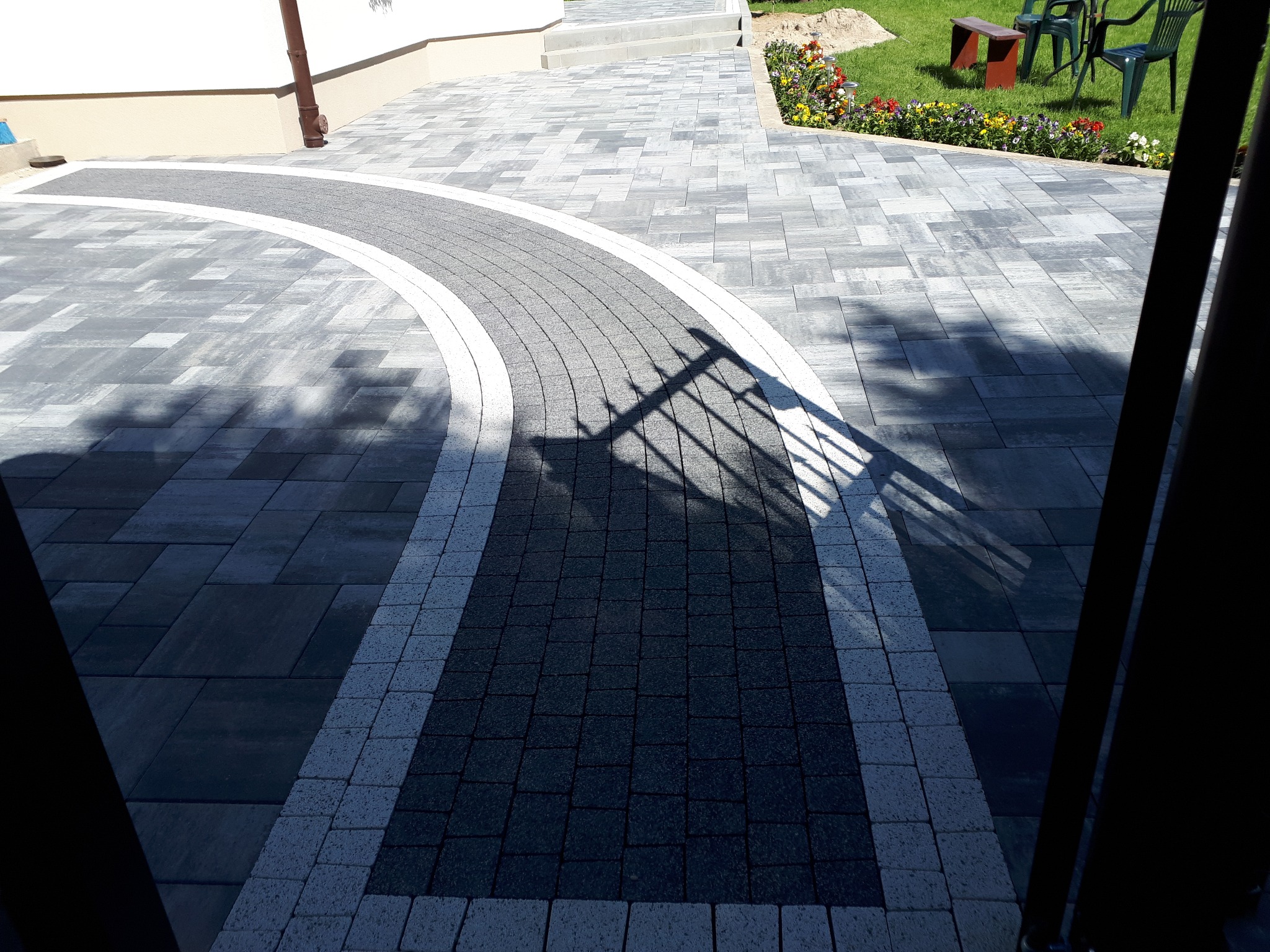 Ścieżka z kostki brukowej w odcieniach szarości, ułożona w łuk, prowadząca do domu z ogrodem w tle, cień bramy na pierwszym planie.