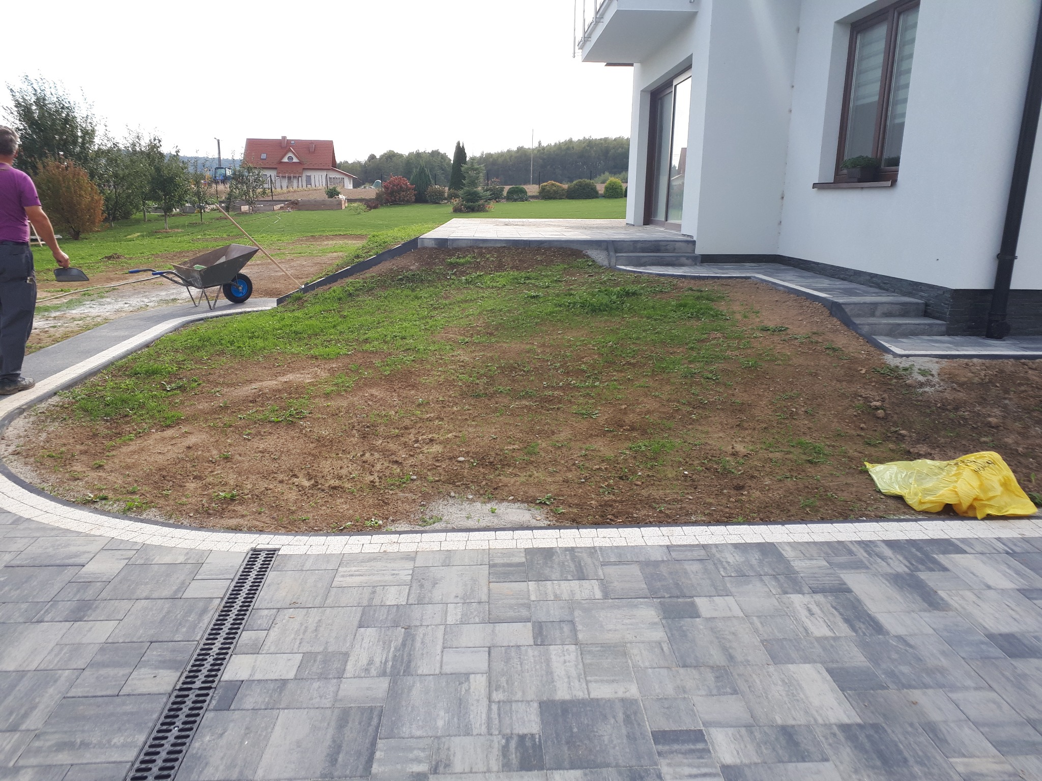 Ułożona szara kostka brukowa z białym obramowaniem i odpływem liniowym, widoczny fragment trawnika przed budynkiem, w tle dom jednorodzinny i pracownik z taczką.