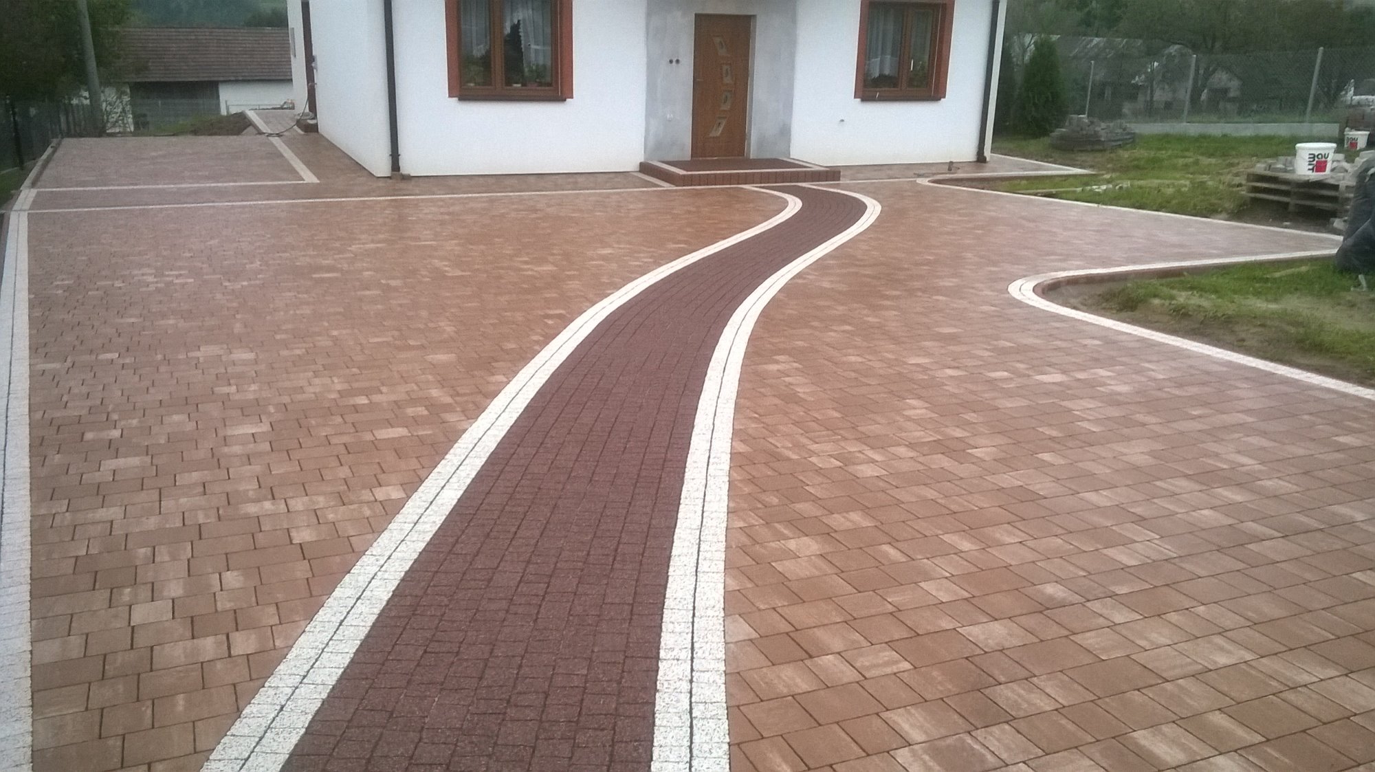 Ułożona kostka brukowa w odcieniach brązu tworząca ścieżkę do domu z białym obramowaniem, widok z góry, jasne oświetlenie.