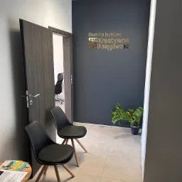 Poczekalnia biura z dwoma czarnymi krzesłami na drewnianych nogach, stolikiem z ulotkami i otwartymi drzwiami do gabinetu z czarnym krzesłem obrotowym w tle. Na ścianie ciemnoszary napis 'Biuro...