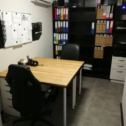 Kreatywna Księgowość sp. z o.o - Wnętrze biura z drewnianym biurkiem, czarnym krzesłem obrotowym, regałem wypełnionym segregatorami i tablicą korkową.