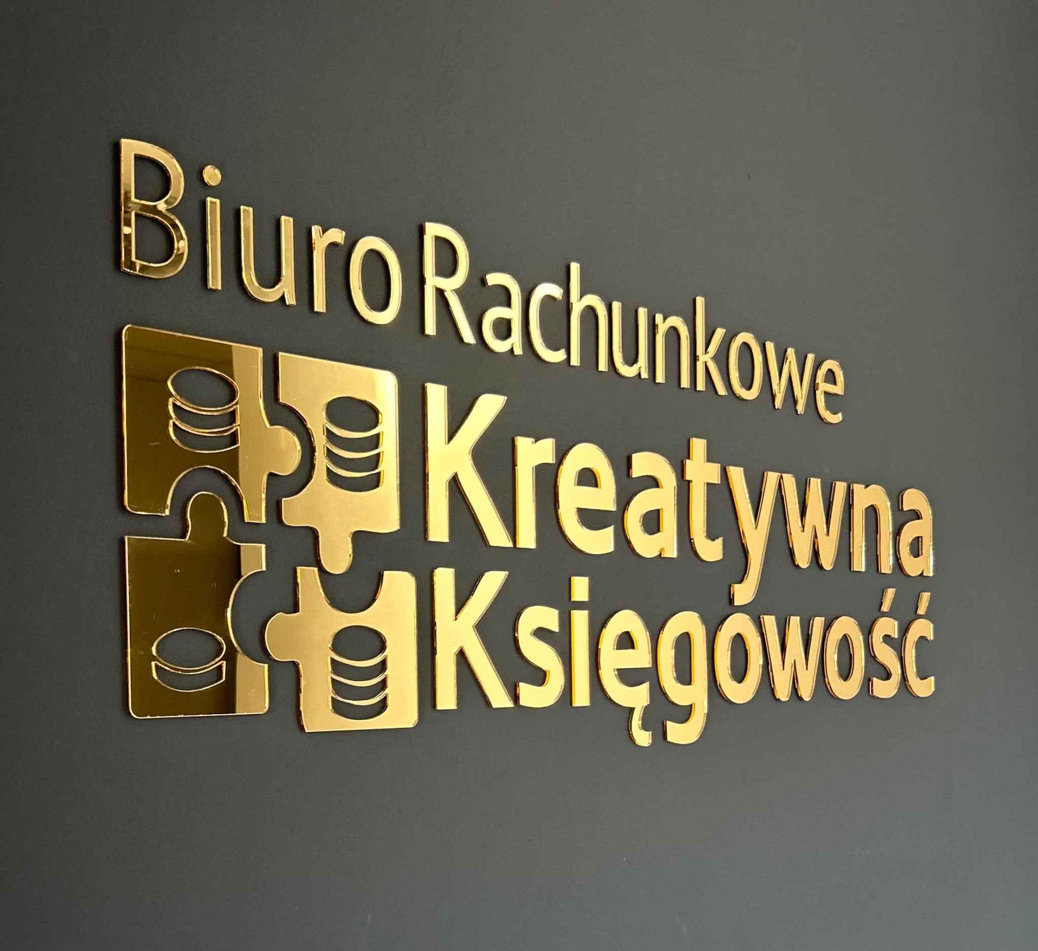Złoty napis 'Biuro Rachunkowe Kreatywna Księgowość' z logo w formie puzzli i stylizowanych monet na ciemnoszarym tle.