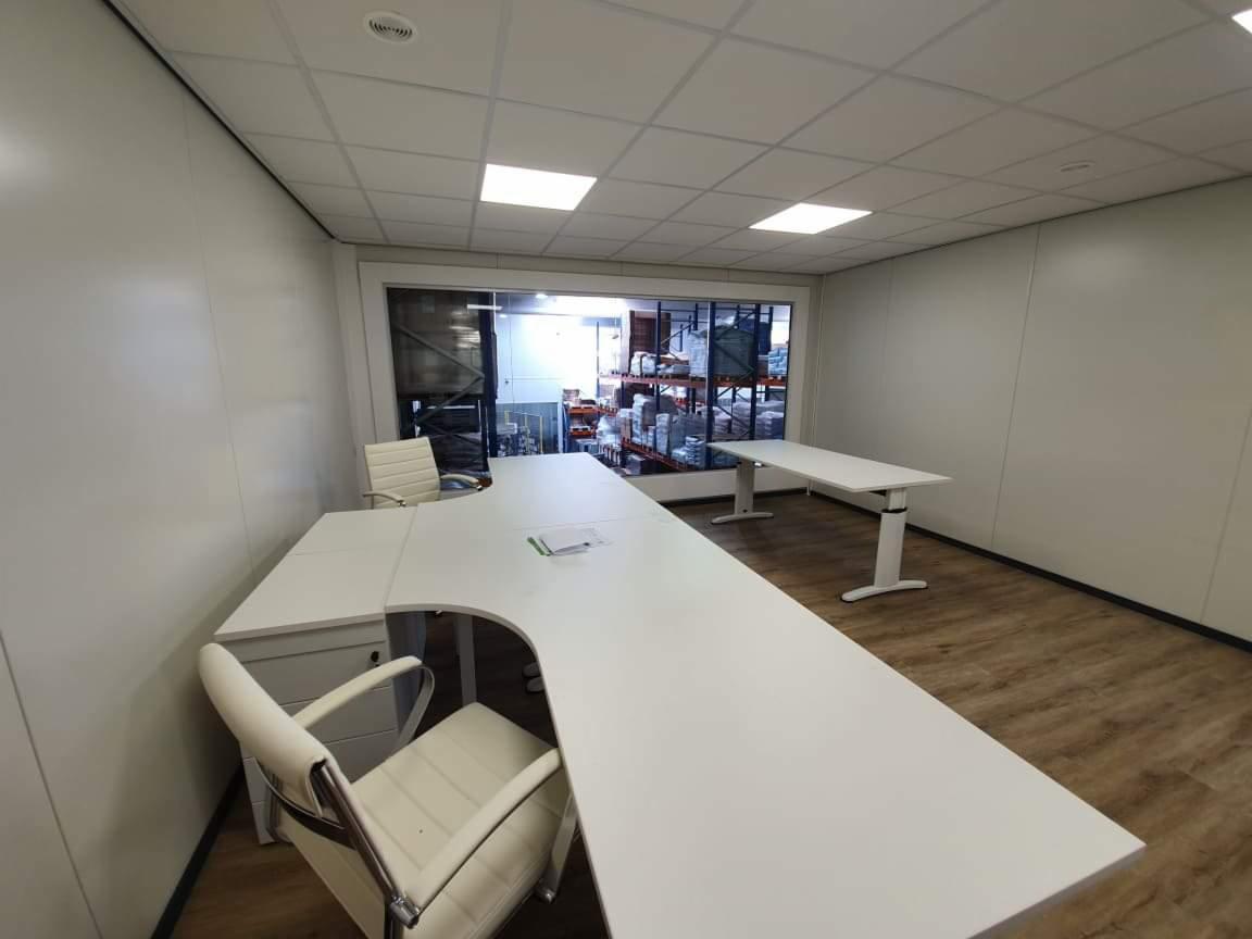 Nowoczesne biuro z białymi ścianami z płyt warstwowych, jasnym drewnianym parkietem i widokiem na magazyn przez szklaną ścianę, wyposażone w białe biurka i krzesła.