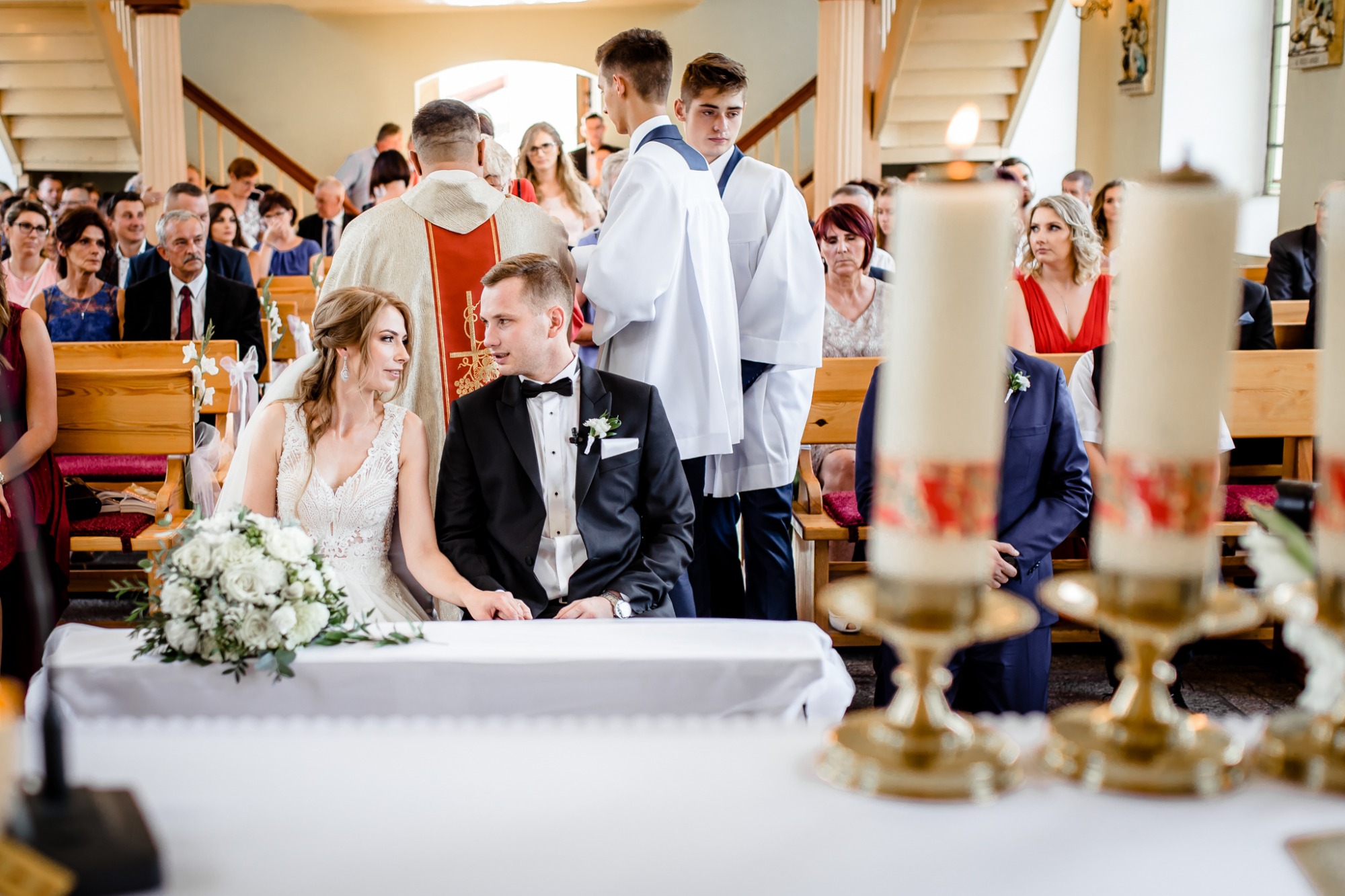 Para młoda siedzi przy stole w kościele podczas ceremonii ślubnej, w tle ksiądz i ministranci, a w pierwszym planie płonące świece.