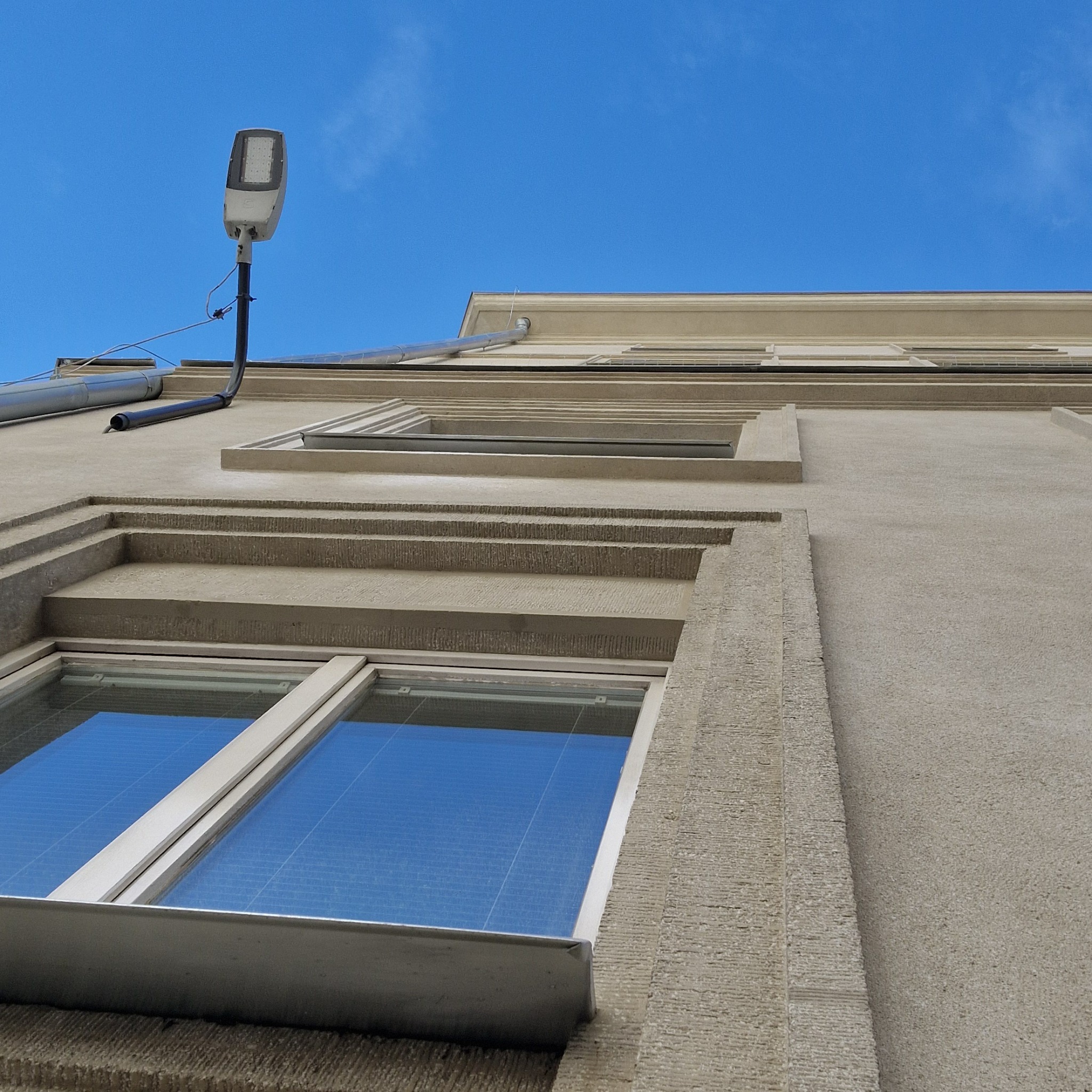 Elewacja kamienicy z oknami, rynną i lampą uliczną na tle błękitnego nieba. Widoczne detale architektoniczne i faktura ściany. Ujęcie z dołu.