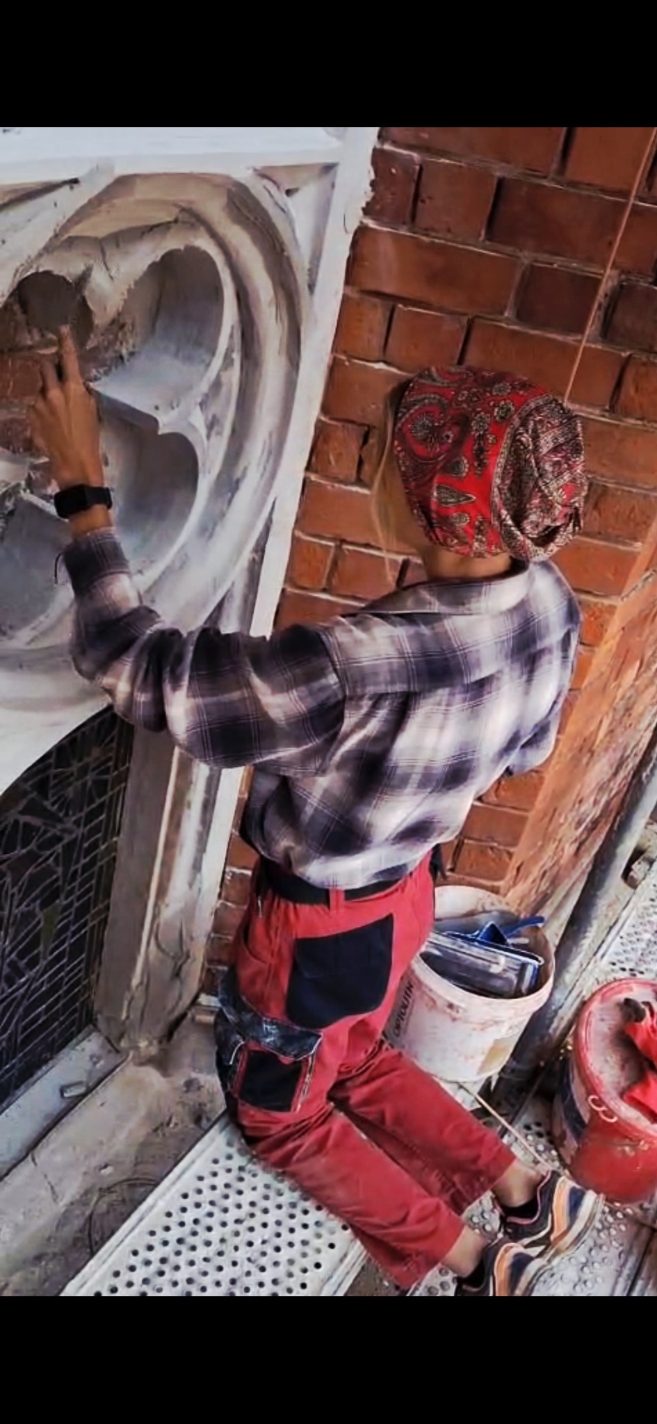 Pracownik w czerwonych spodniach i kraciastej koszuli naprawia fragment zabytkowej fasady z cegły, stojąc na rusztowaniu. Widoczne narzędzia i wiadro z zaprawą.