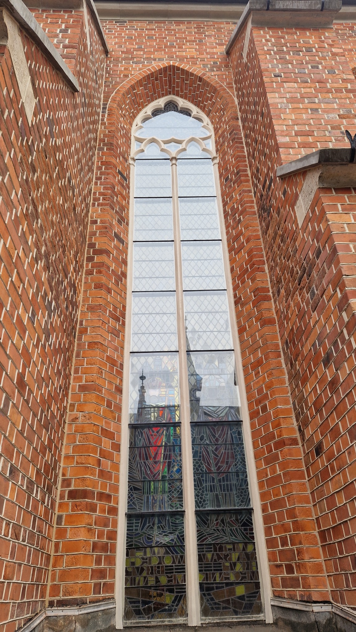 Gotyckie okno z witrażem w ceglanej fasadzie kościoła, sfotografowane z dołu. Witraż odbija fragment wieży. Charakterystyczna architektura sakralna.