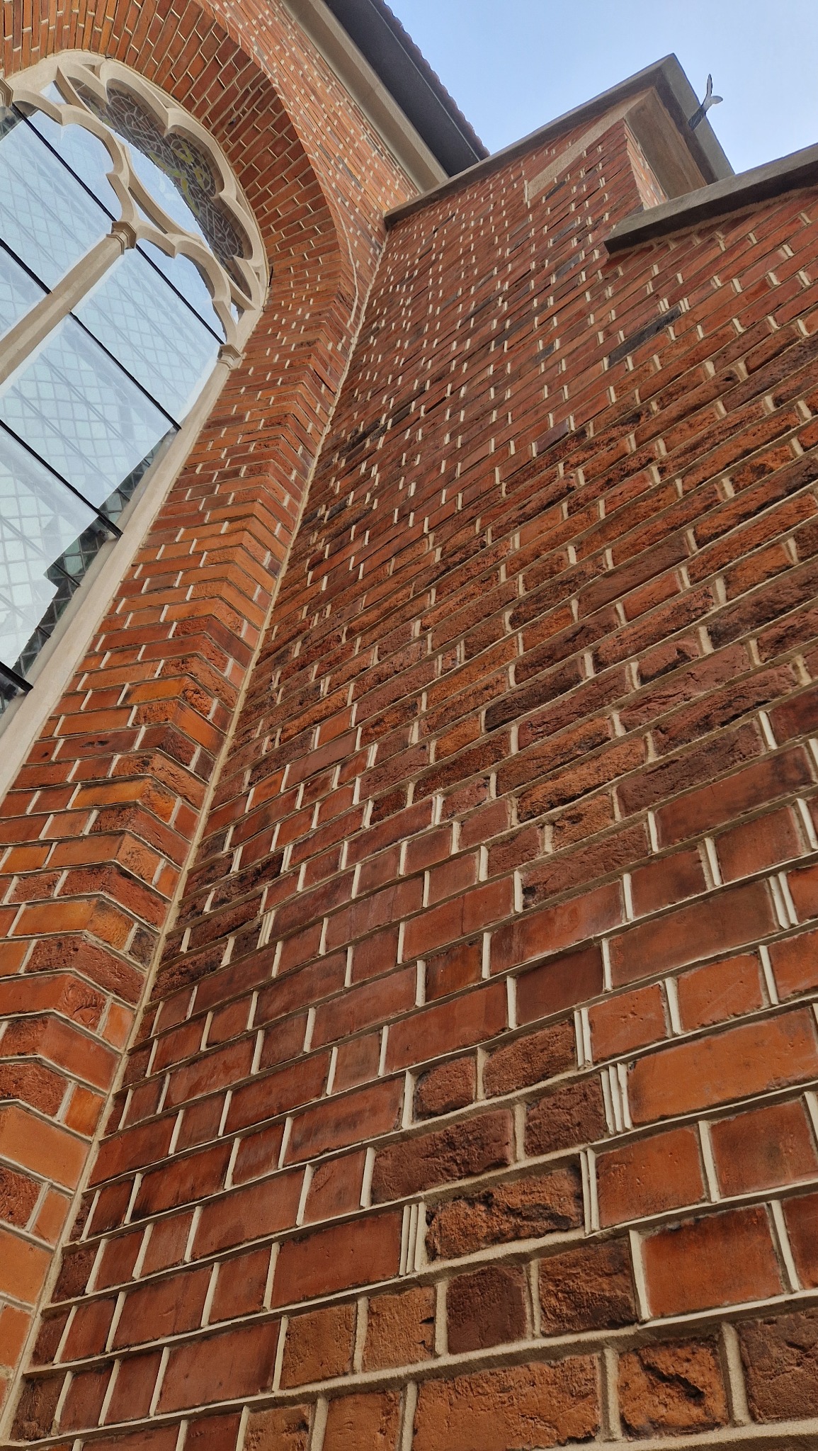 Ceglana ściana budynku z oknem w stylu gotyckim, widziana pod kątem z dołu, eksponująca detale muru i architekturę zabytkową.