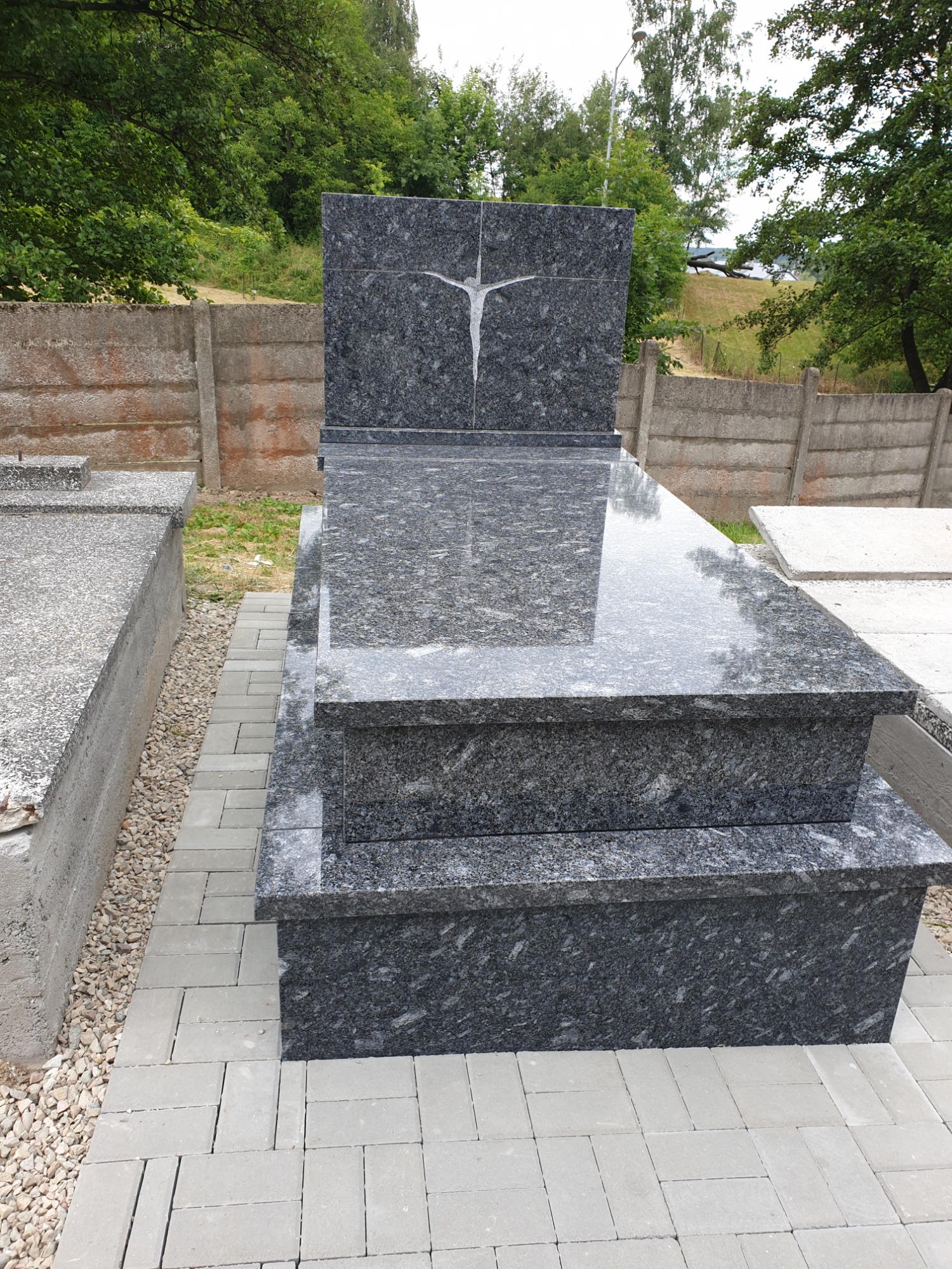 Polerowany, dwupoziomowy pomnik z ciemnego granitu z wyrytym motywem figury na pionowej płycie, otoczony brukowaną ścieżką i betonowym ogrodzeniem.