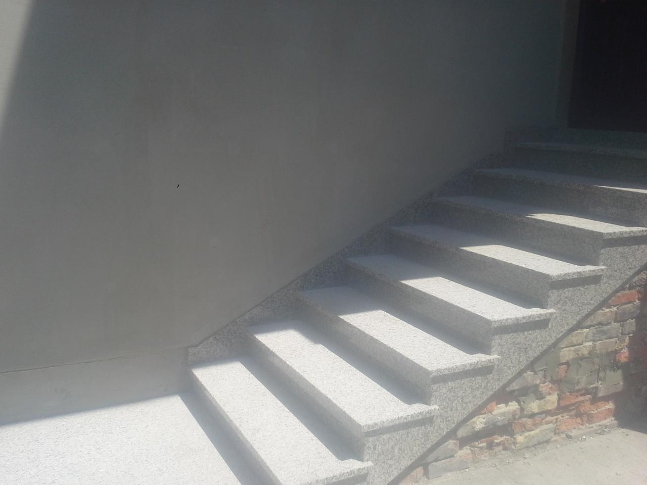 Zewnętrzne schody z jasnego, nakrapianego kamienia prowadzące do ciemnego wejścia, z fragmentem ceglanej ściany u podstawy.