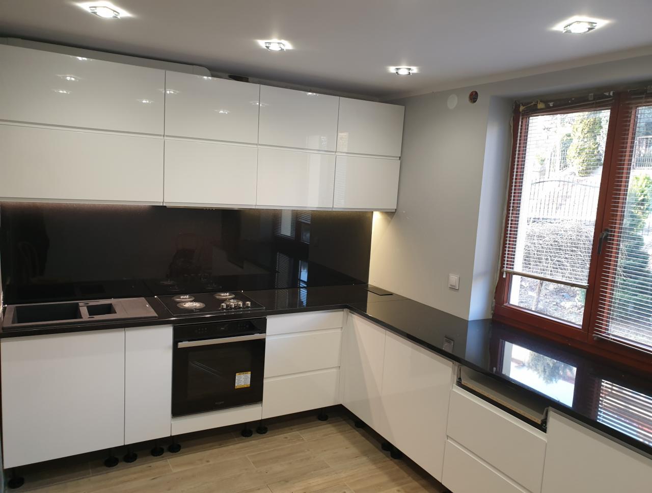 Nowoczesna kuchnia z białymi szafkami i czarnym blatem, widok na okno z żaluzjami.