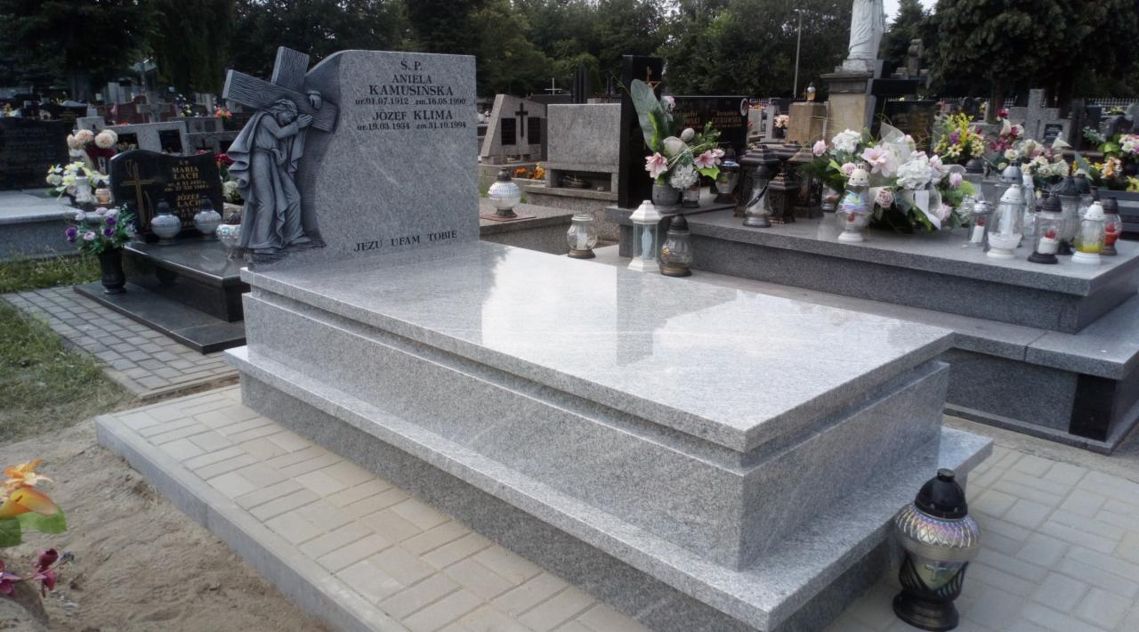 Jasnoszary, granitowy pomnik z rzeźbą Chrystusa niosącego krzyż, z inskrypcjami imion i dat, otoczony lampionami i kwiatami na cmentarzu.