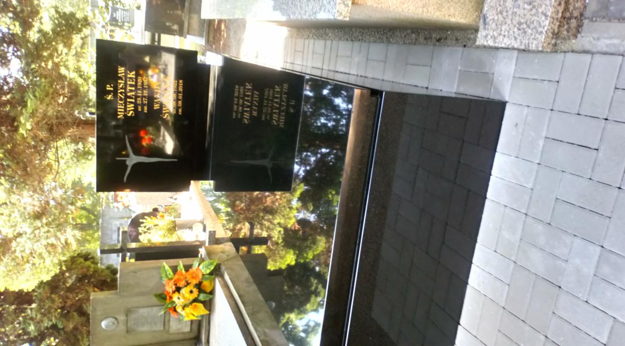 Czarny, wypolerowany pomnik z jasnym, geometrycznym symbolem i inskrypcją, otoczony szarą kostką brukową i kwiatami w odcieniach żółci i pomarańczy.