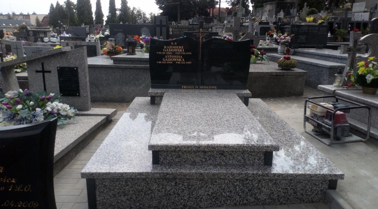 Granitowy pomnik nagrobny z ciemną tablicą inskrypcyjną i złotym krzyżem, otoczony innymi nagrobkami na cmentarzu, z generatorem w tle.