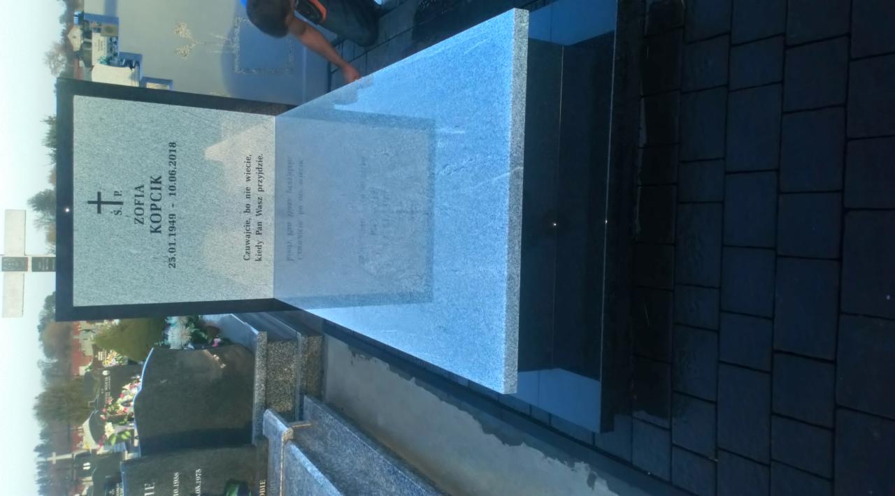 Jasnoszary nagrobek z ciemną obwódką i inskrypcją, usytuowany na ciemnej kostce brukowej, widoczny fragment osoby podczas montażu.