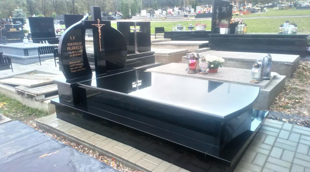 Czarny, polerowany pomnik z krzyżem i złotym wizerunkiem Chrystusa na cmentarzu, z inskrypcjami na bocznej płycie i kwiatami na sąsiednim grobie.