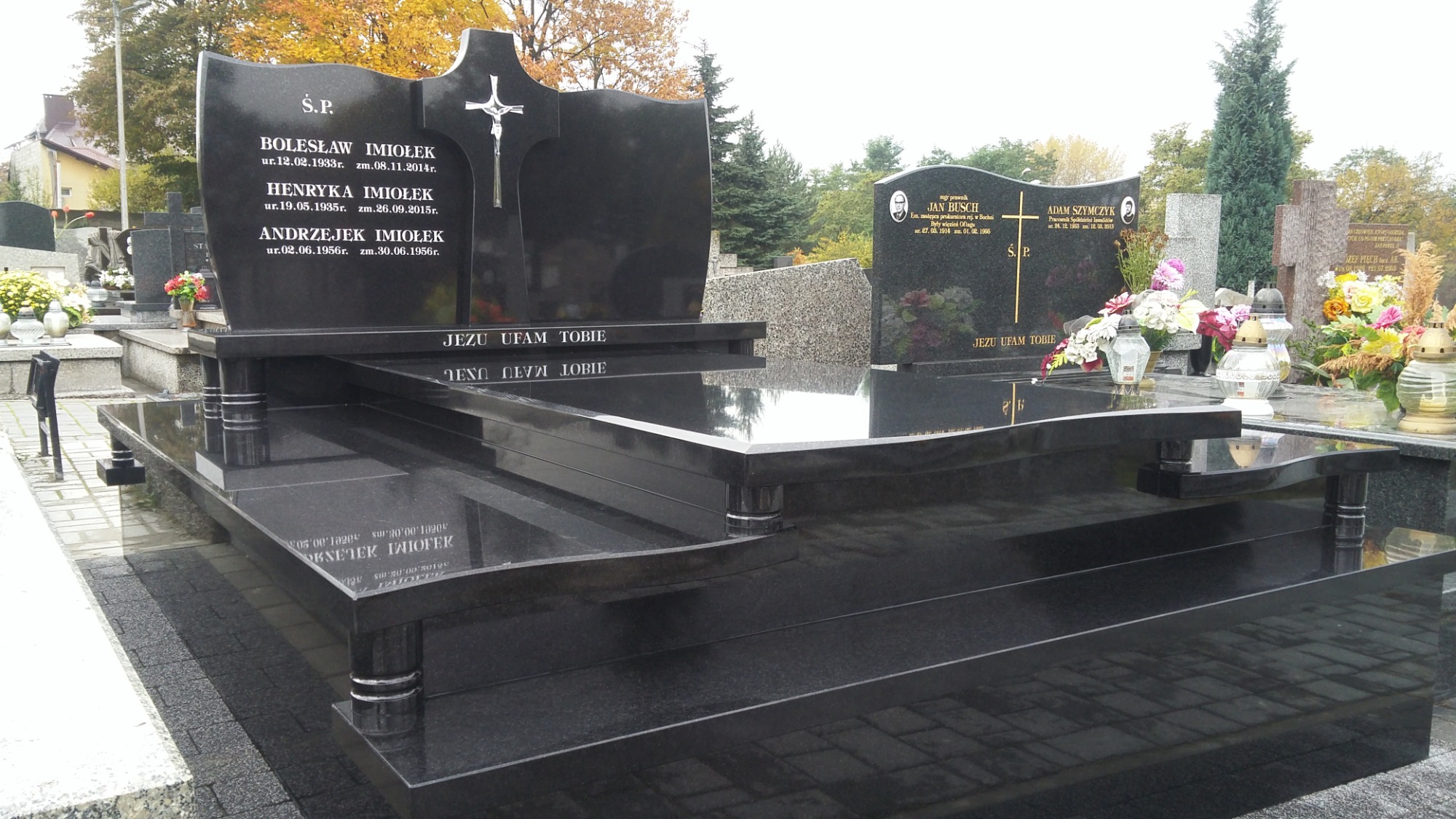 Czarny, wielopoziomowy pomnik nagrobny z inskrypcjami i krzyżem, otoczony innymi nagrobkami na cmentarzu, widoczne jesienne drzewa w tle.