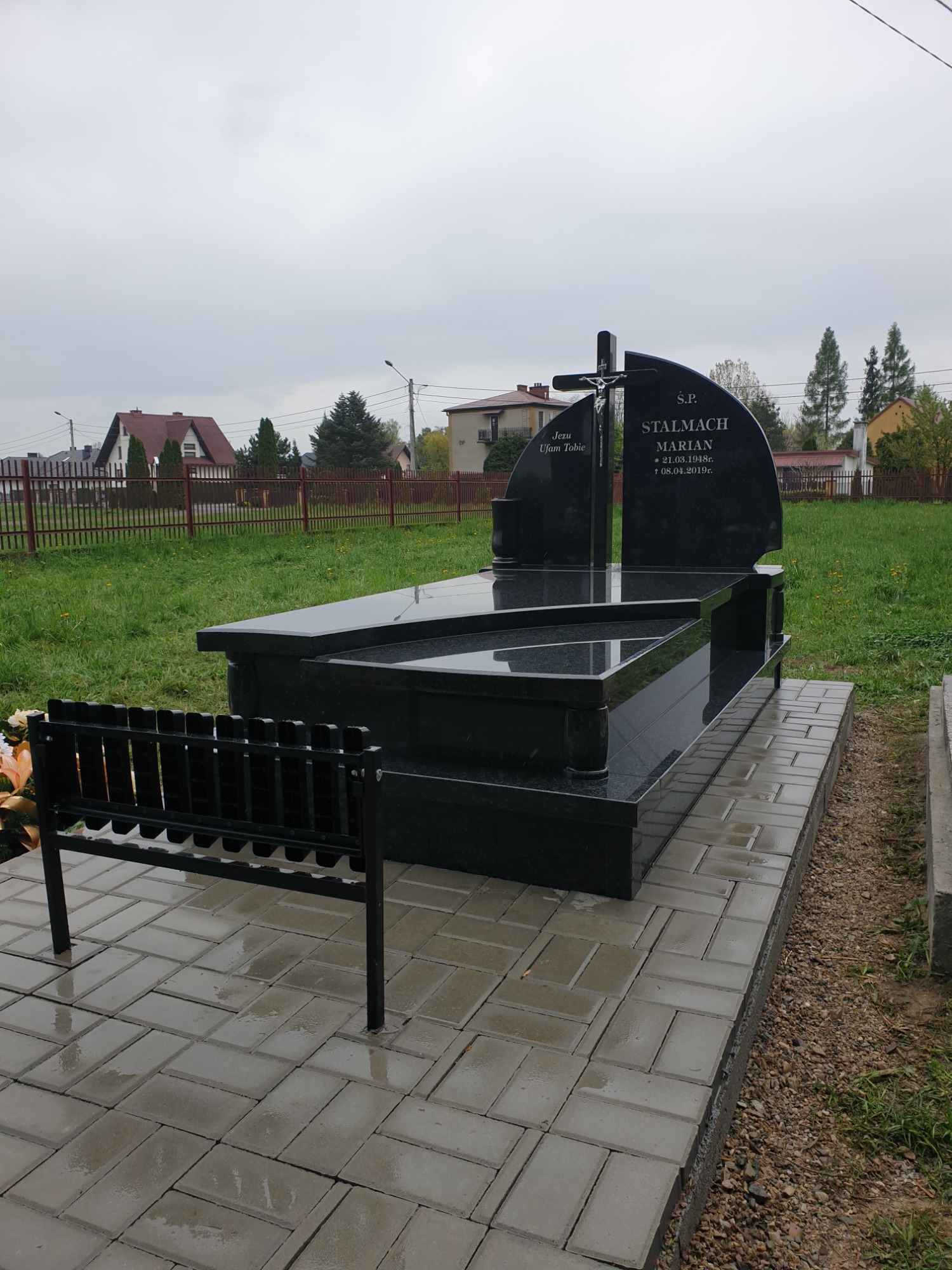 Czarny, błyszczący pomnik z wygrawerowanym napisem i krzyżem, ustawiony na wyłożonym kostką brukową podeście. Obok pomnika znajduje się metalowa ławeczka.
