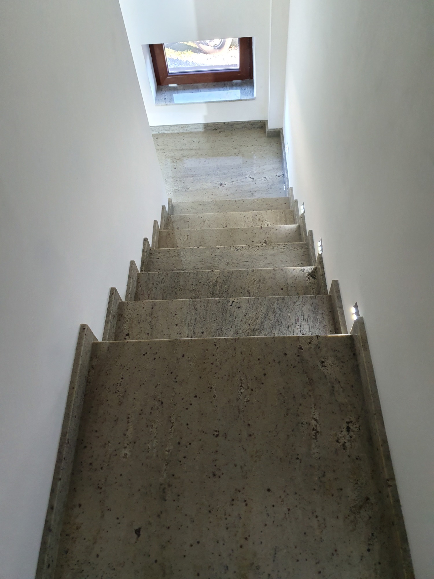 Schody wewnętrzne z szarego kamienia z delikatnym wzorem, oświetlone małymi lampkami LED w ścianie, prowadzące do okna dachowego.
