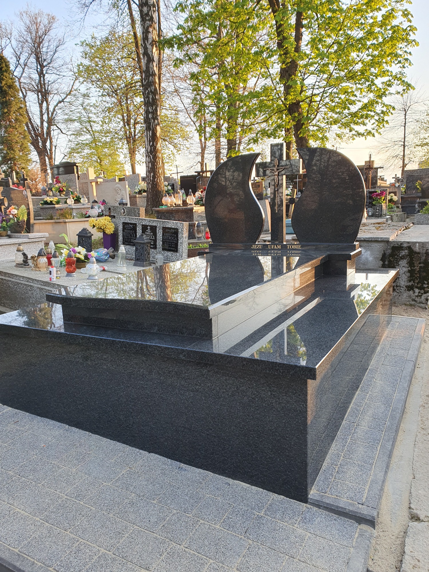 Nowoczesny, czarny pomnik z dwoma łukowatymi płytami i krzyżem, umieszczony na cmentarzu w słoneczny dzień, z widocznymi innymi grobami w tle.