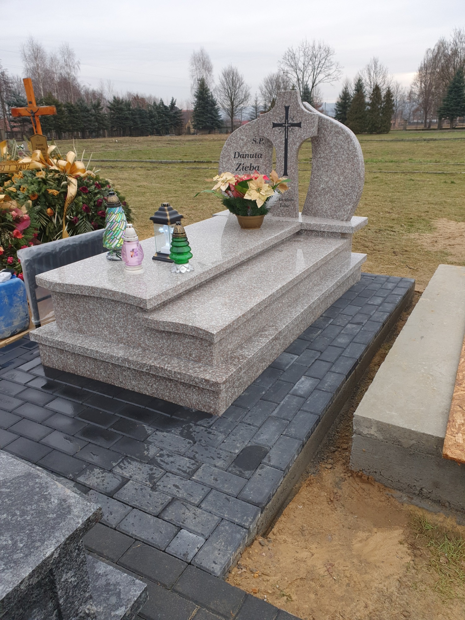 Granitowy nagrobek z ciemnoszarą kostką brukową u podstawy, ozdobiony kwiatami i zniczami, na tle cmentarza w pochmurny dzień.
