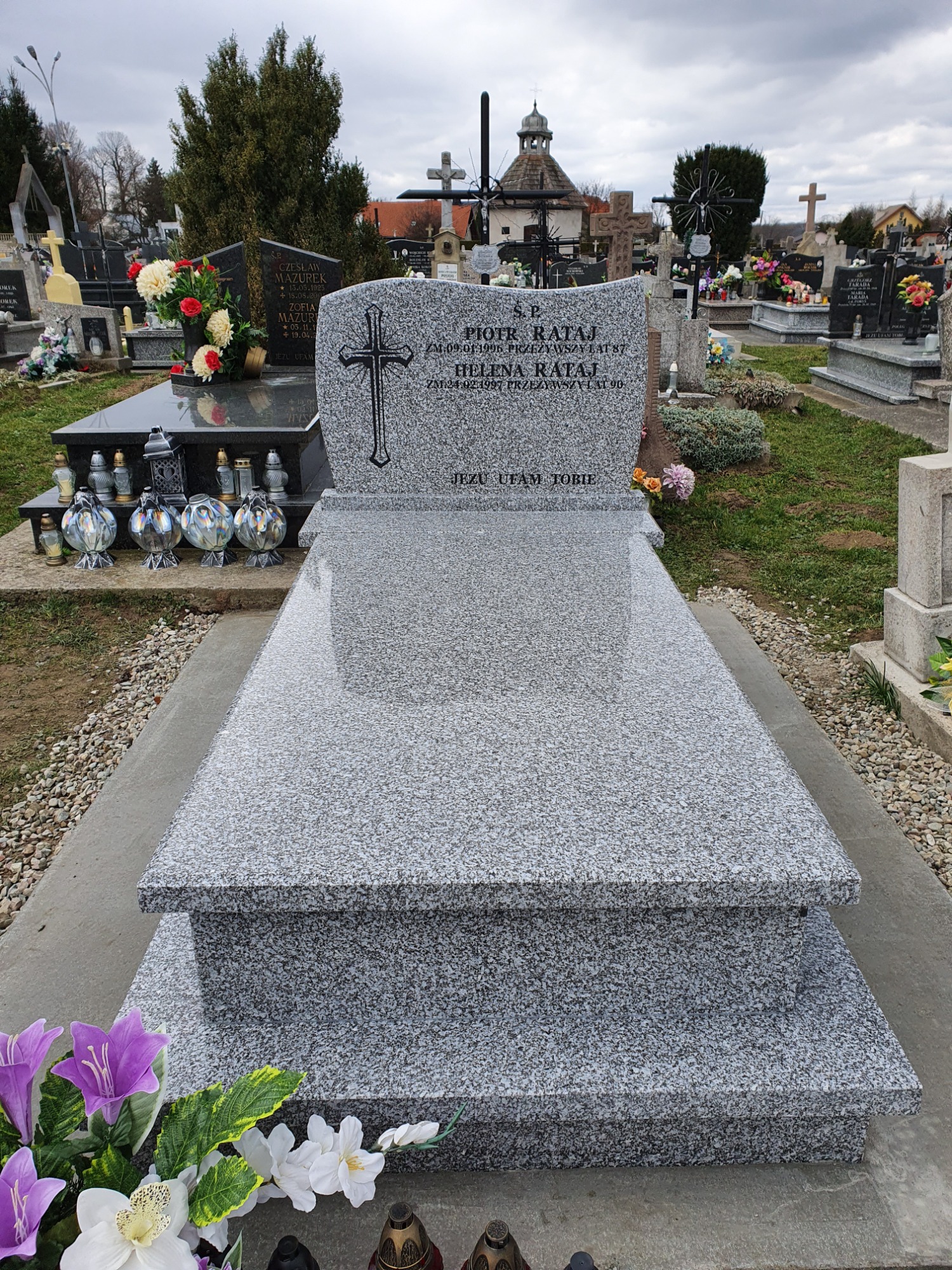 Jasnoszary, dwupoziomowy pomnik z wygrawerowanym krzyżem i inskrypcją na cmentarzu, otoczony kwiatami i zniczami, z widocznym w tle kościołem.