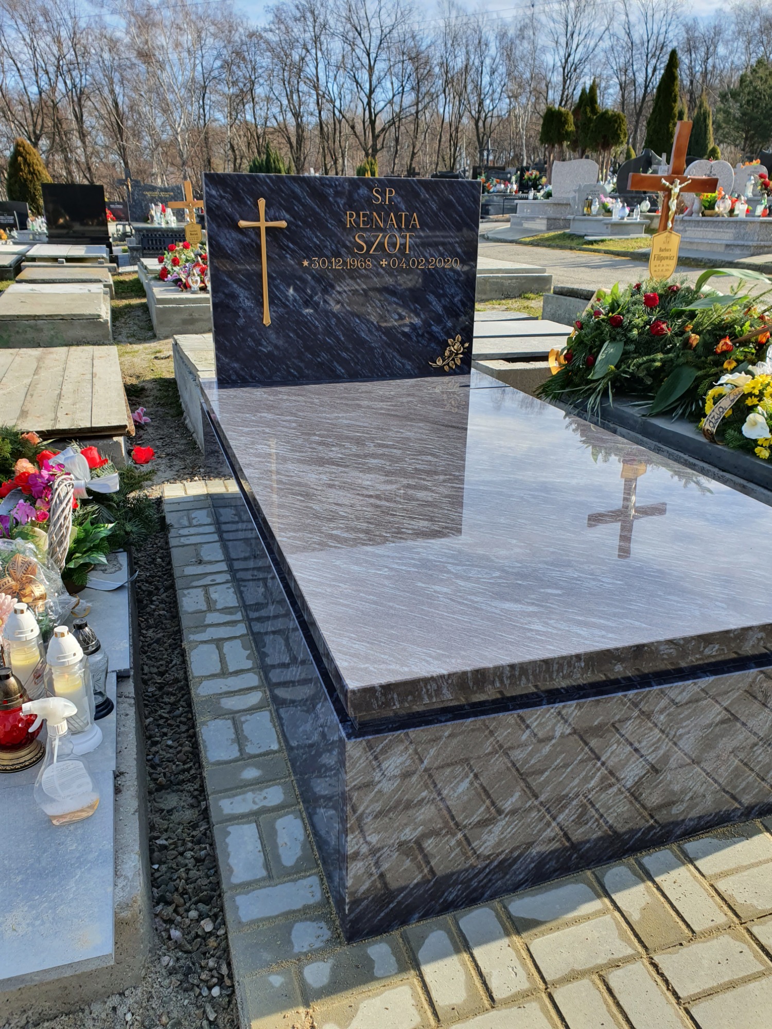 Polerowany nagrobek z ciemnego granitu z wygrawerowanym złotym krzyżem i inskrypcją na cmentarzu, z refleksami na powierzchni i kompozycjami kwiatowymi w tle.