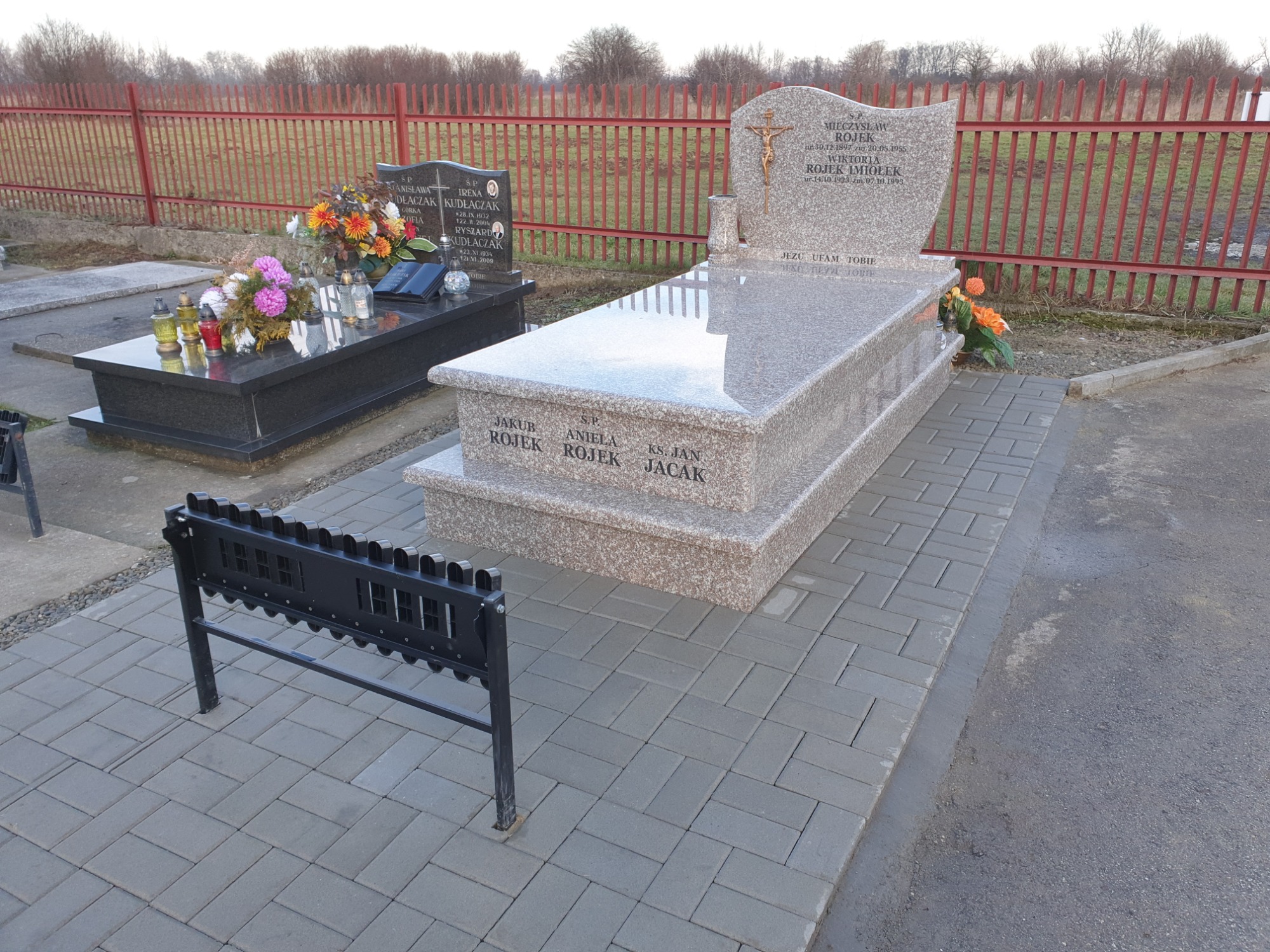 Jasny, granitowy nagrobek z inskrypcjami i krzyżem, otoczony kostką brukową, na cmentarzu z widocznym ogrodzeniem i innymi nagrobkami w tle.