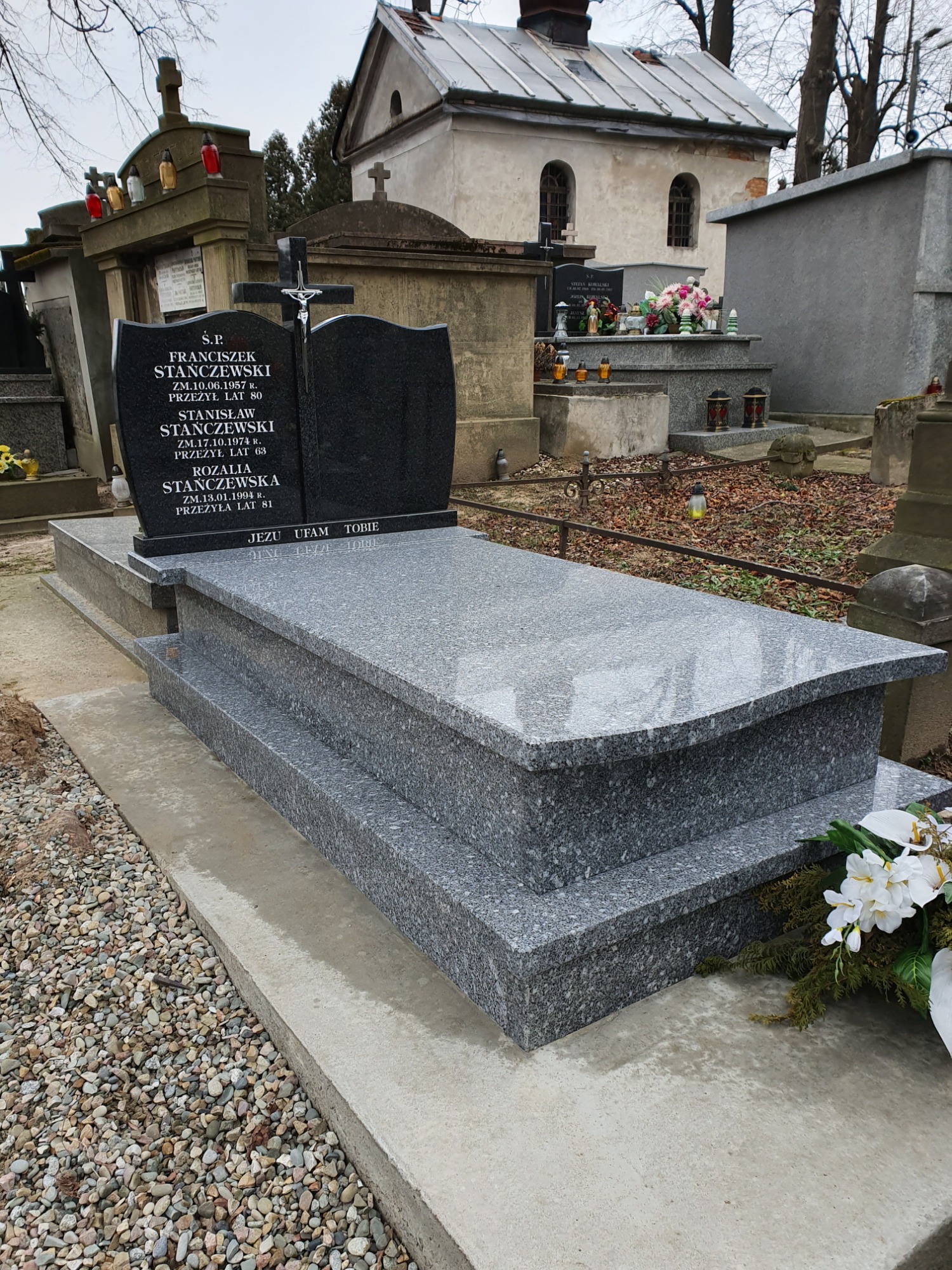 Szary, granitowy pomnik nagrobny z wygrawerowanymi danymi zmarłych na tle cmentarza z innymi grobami i kaplicą.