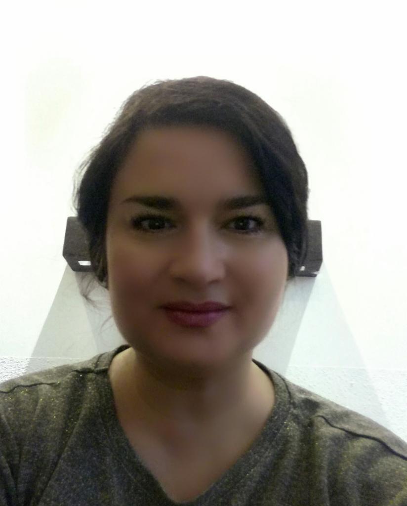 Portret uśmiechniętej kobiety z ciemnymi włosami, ubrana w szarą bluzkę z delikatnymi złotymi akcentami, na tle białej ściany z elementami oświetlenia.