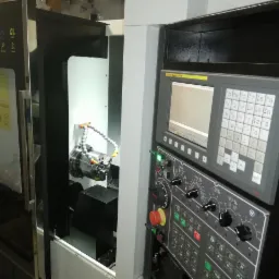 Automatyczna tokarka CNC w trakcie obróbki detalu, widoczny panel sterowania z ekranem i przyciskami, chłodziwo skierowane na obrabiany element.