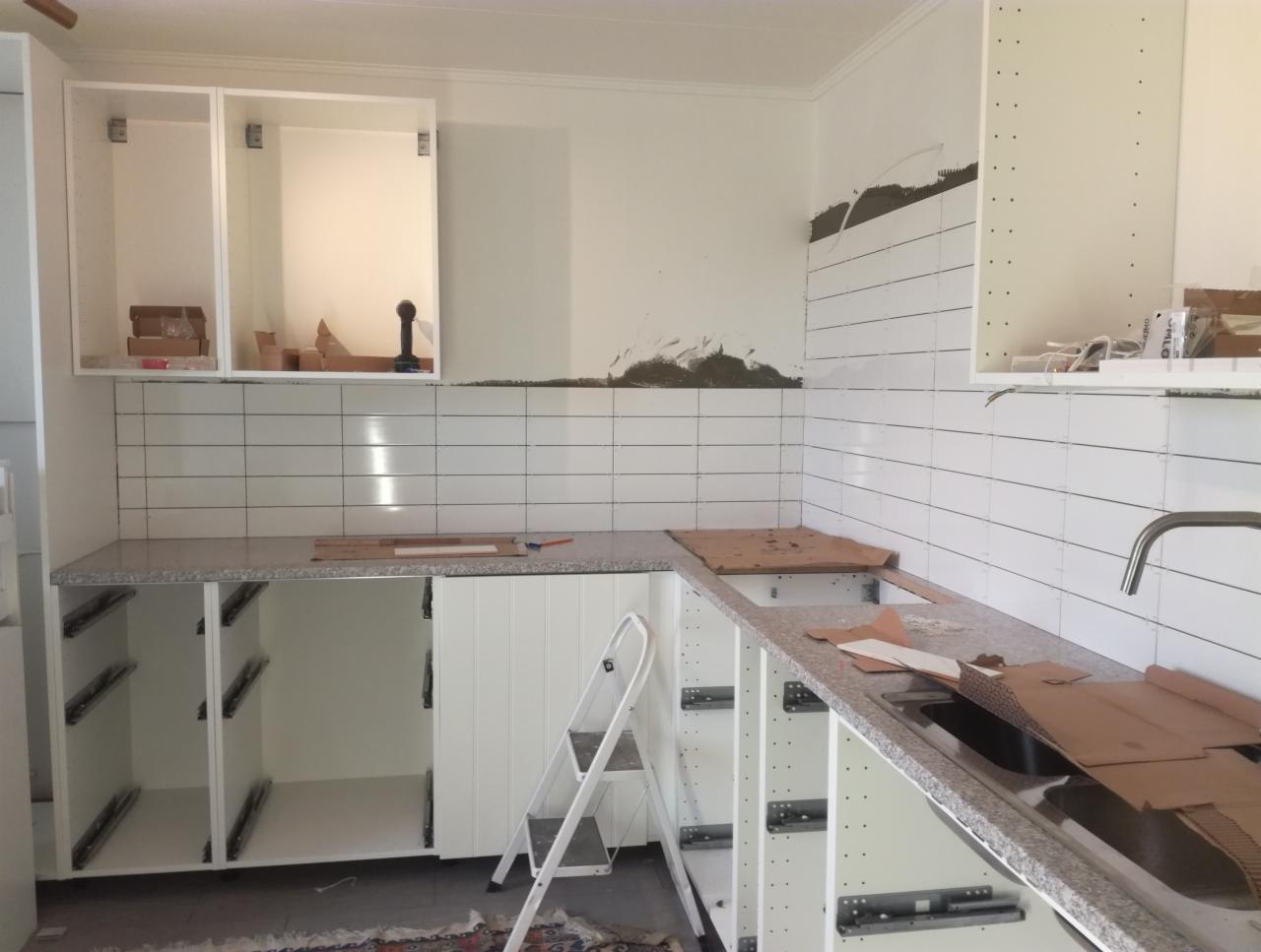 Kuchnia w trakcie montażu: białe płytki na ścianie, częściowo ułożone, białe szafki kuchenne bez frontów, blat z jasnego kamienia, drabina oparta o szafkę.