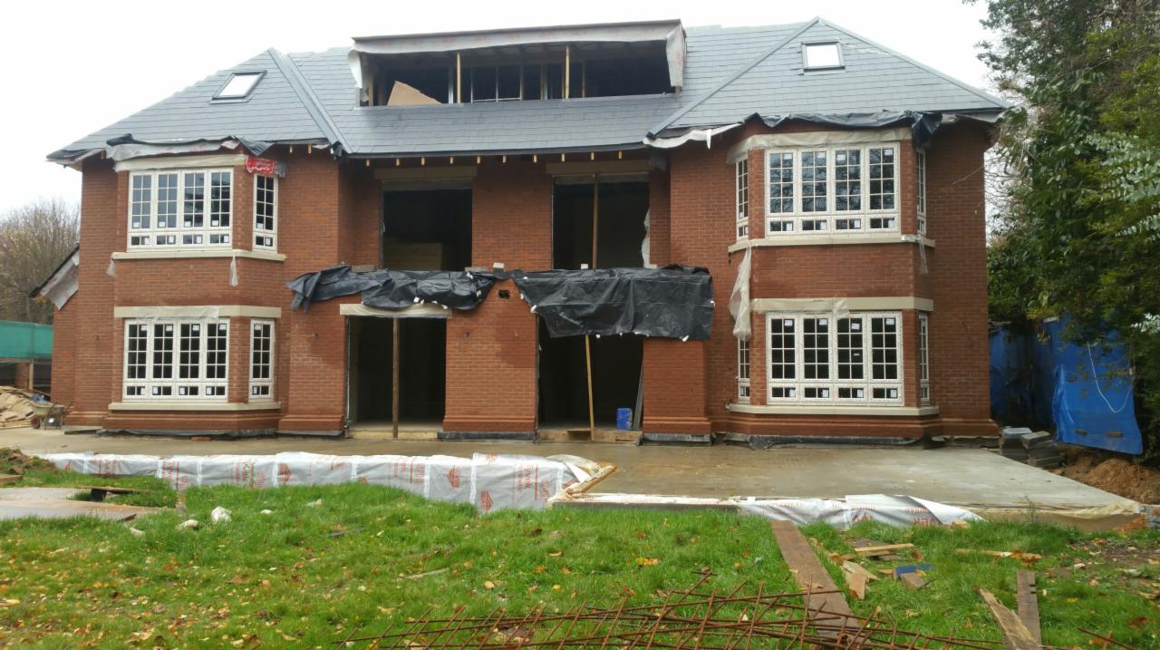 Dwupiętrowy dom z czerwonej cegły w trakcie budowy, z widocznymi oknami z białymi ramami, szarym dachem i niezabezpieczonymi otworami okiennymi i drzwiowymi, z fragmentami folii ochronnej...