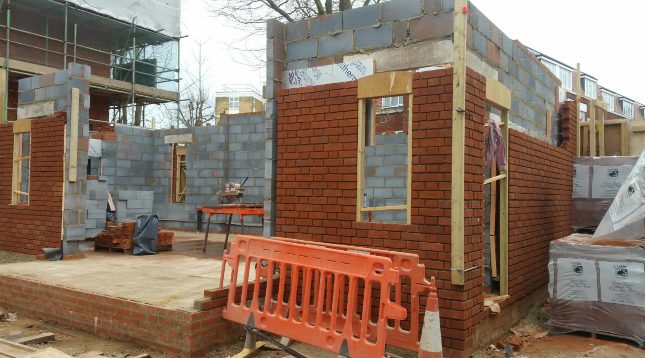 Budowa domu: murowane ściany z czerwonej cegły i szarego bloczka, widoczne otwory okienne z drewnianą ramą, na pierwszym planie pomarańczowe bariery ochronne.