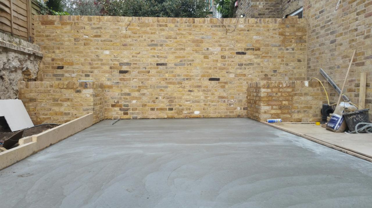 Świeżo wylana betonowa posadzka na tle ceglanej ściany z żółtej cegły z widocznymi narzędziami murarskimi i materiałami budowlanymi.