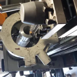 Metalowy cylinder w uchwycie tokarki podczas obróbki. Widoczny nóż tokarski i fragment sterownika numerycznego.