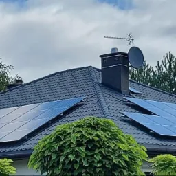 Panele słoneczne na grafitowym dachu domu jednorodzinnego z anteną satelitarną i kominem w tle, otoczone zielenią.