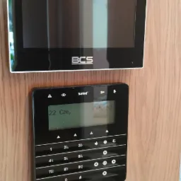 Panel sterowania alarmem firmy Satel z wyświetlaczem i klawiaturą numeryczną, zamontowany obok wideodomofonu BCS na ścianie o strukturze drewna.