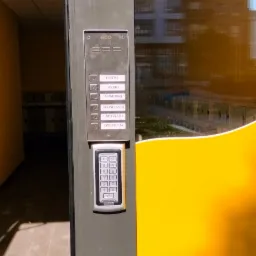 Panel wejściowy z domofonem i klawiaturą numeryczną, zamontowany na szarej ramie drzwi, z widocznymi przyciskami nazw lokatorów: Listki, Rybki, Chmurki, Słoneczka, Motylki, Dyrekcja.